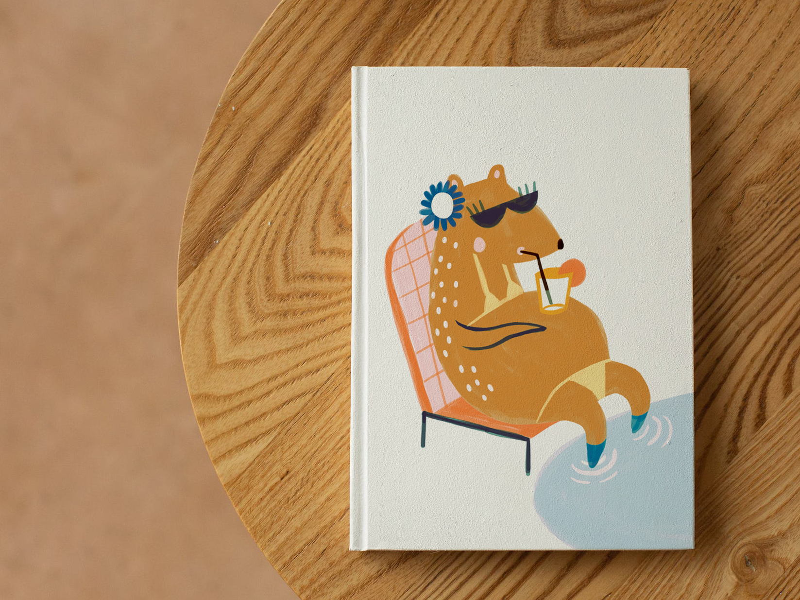 livre fermé et posé sur une table en bois. Il n'y a pas de titre, juste une illustration enfantine d'un ours buvant à la paille, une limonade posée sur son gros ventre. Il a les pattes dans l'eau, une fleur sous l'oreille. Il se prélasse sur sa chaise de plage